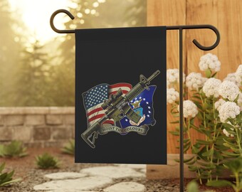 Puede incluir: Bandera de jardín negra con una ilustración de un rifle, la bandera estadounidense y el emblema de la Fuerza Aérea de los EE. UU. La bandera tiene el texto "HOIST THE COLOURS" en una pancarta. La bandera se muestra en un soporte de metal negro.