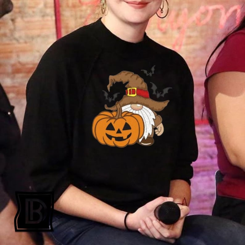 May include: A black long-sleeve sweatshirt featuring a graphic print of a gnome wearing a brown witch hat with a red band, holding a large orange jack-o'-lantern pumpkin. Several small black bats are flying around the gnome's hat.