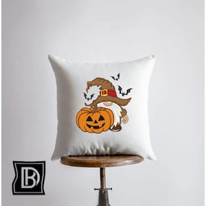 May include: A square white throw pillow featuring a graphic illustration of a gnome wearing a brown witch hat with a red band, holding a large orange jack-o'-lantern pumpkin. Several small black bats are flying around the gnome's hat.