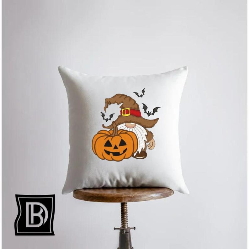 May include: A square white throw pillow featuring a graphic illustration of a gnome wearing a brown witch hat with a red band, holding a large orange jack-o'-lantern pumpkin. Several small black bats are flying around the gnome's hat.