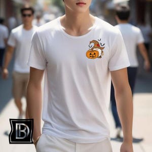 May include: A white short-sleeved t-shirt featuring a small graphic on the left chest area. The graphic depicts a gnome wearing a brown witch hat, standing next to a bright orange jack-o'-lantern pumpkin, surrounded by small black bats.