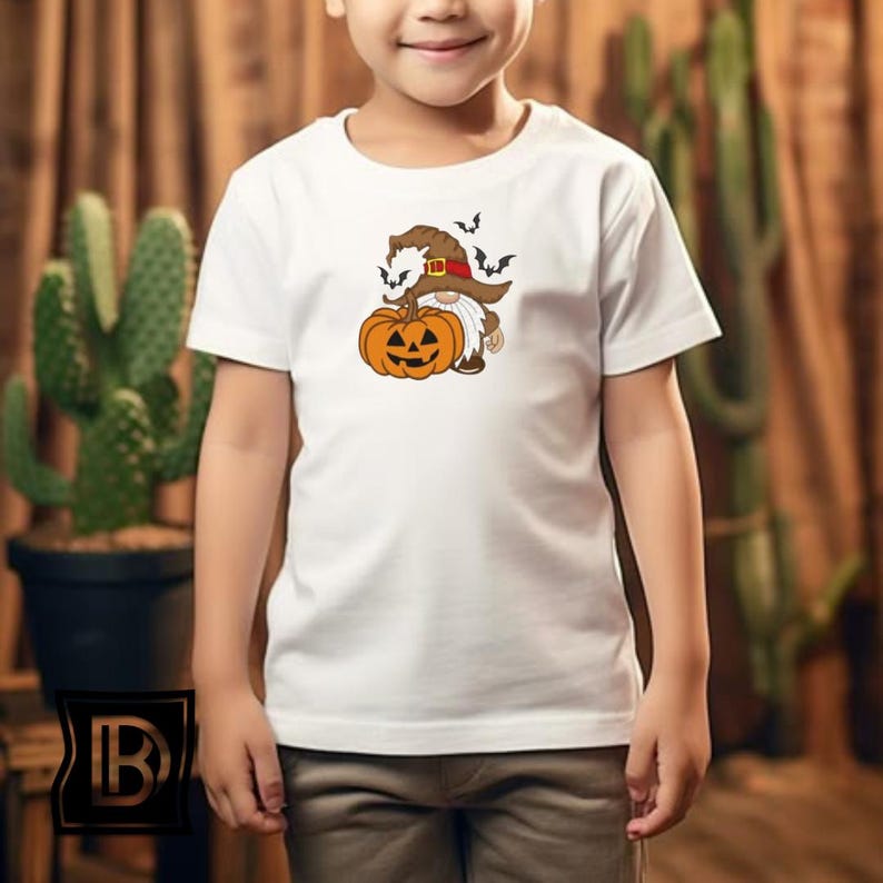 May include: A white short-sleeved t-shirt featuring a graphic of a gnome wearing a brown witch hat and holding a bright orange jack-o'-lantern pumpkin. Small black bats are illustrated flying above the gnome's hat on the center of the chest.