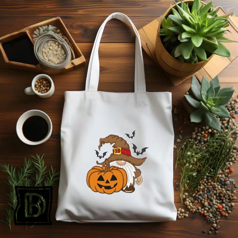 May include: A white tote bag featuring a graphic print of a gnome wearing a brown witch hat with a red band, standing next to an orange jack-o'-lantern pumpkin. Several small black bats are flying around the gnome's hat on the bag's surface.