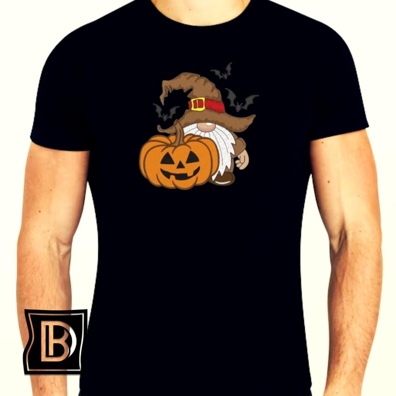May include: A black short-sleeved t-shirt featuring a graphic print of a gnome wearing a brown witch hat with a red band, holding an orange jack-o'-lantern pumpkin. Several small black bats are flying around the gnome's hat on the chest area.