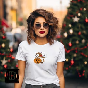 May include: A white short-sleeved t-shirt featuring a small graphic print of a gnome wearing a brown witch hat and holding an orange jack-o'-lantern pumpkin, with three small black bats flying above the gnome's hat on the chest area.