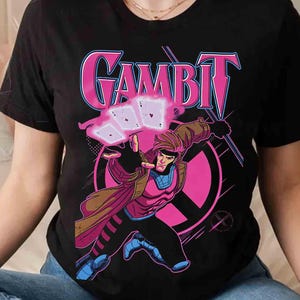 Pode incluir: Camiseta preta com um gráfico do personagem Gambit da Marvel Comics. O design inclui a palavra "GAMBIT" em rosa, um fundo circular rosa e uma ilustração de Gambit jogando cartas.