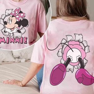 May include: Pink t-shirt featuring a Minnie Mouse graphic. The front of the shirt shows Minnie bursting through a torn paper effect, with the name "MINNIE" below. The back of the shirt has a matching design of Minnie's feet and bow.