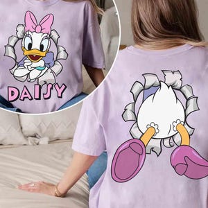 Puede incluir: Camiseta lila claro con un dise&ntilde;o de Daisy Duck. La parte delantera muestra a Daisy Duck saliendo de un agujero rasgado, con el nombre "DAISY" debajo. La parte trasera de la camiseta muestra la parte trasera y los pies de Daisy.