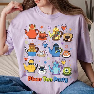 May include: A light purple t-shirt featuring a colorful graphic of cartoon teapots and teacups. The phrase "Pixar Tea Party" is printed in orange and blue. The design includes stars and balloons.