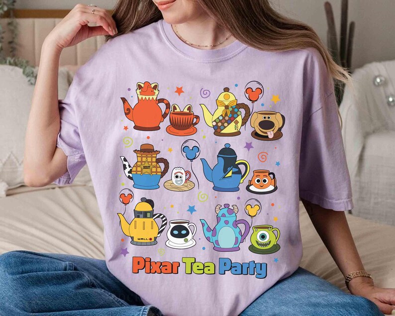 May include: A light purple t-shirt featuring a colorful graphic of cartoon teapots and teacups. The phrase "Pixar Tea Party" is printed in orange and blue. The design includes stars and balloons.