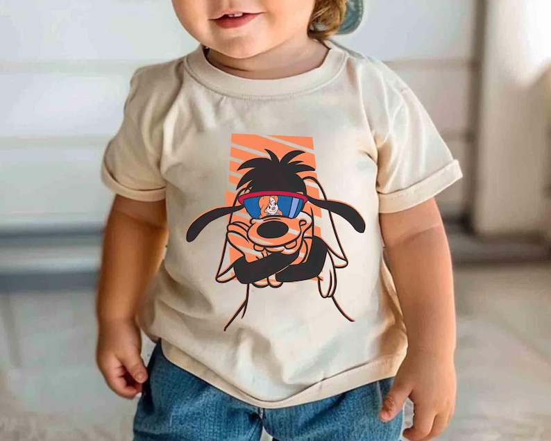 May include: A toddler wearing a beige t-shirt with a cartoon dog wearing sunglasses and a backwards baseball cap. The dog is wearing a red and blue bandana around its neck.