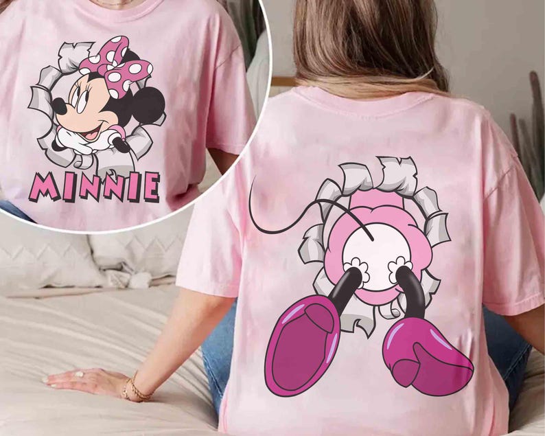 May include: Pink t-shirt featuring a Minnie Mouse graphic. The front of the shirt shows Minnie bursting through a torn paper effect, with the name "MINNIE" below. The back of the shirt has a matching design of Minnie's feet and bow.