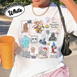 May include: White t-shirt with a colorful Disney's Hollywood Studios graphic. The design includes cartoon characters, movie-themed elements, and the year 2026. The shirt has the words "Comfort Colors" and "White" in a circular design.