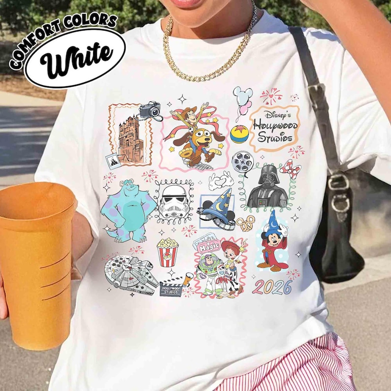May include: White t-shirt with a colorful Disney's Hollywood Studios graphic. The design includes cartoon characters, movie-themed elements, and the year 2026. The shirt has the words "Comfort Colors" and "White" in a circular design.