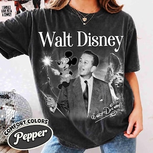 Puede incluir: Una camiseta de manga corta gris carb&oacute;n con un estampado gr&aacute;fico de Walt Disney en traje y corbata, junto a un dibujo de Mickey Mouse sosteniendo una varita. El texto Walt Disney est&aacute; impreso en blanco sobre la imagen, con una firma debajo.