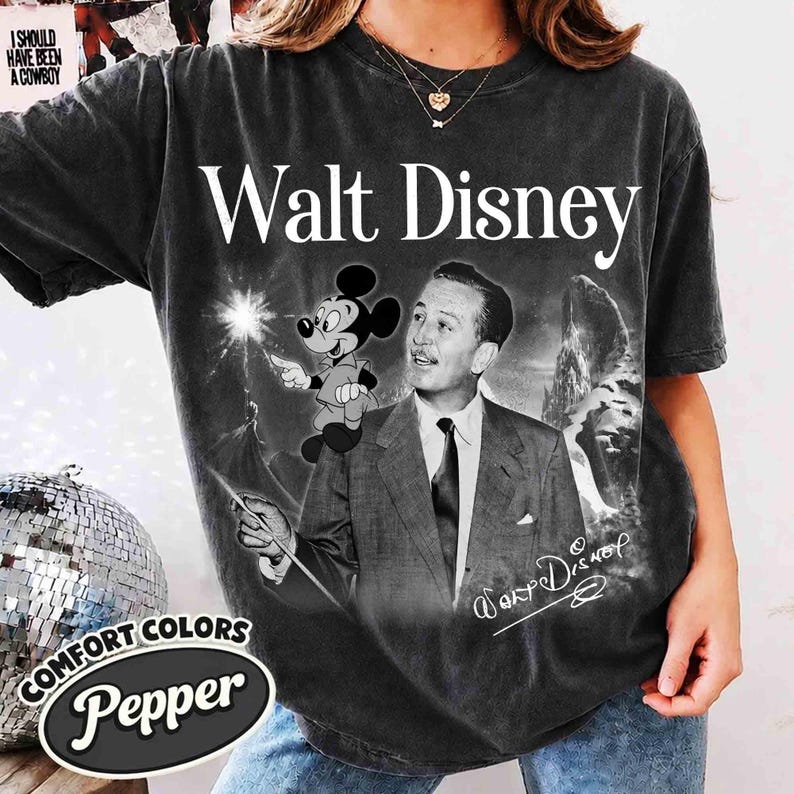 Puede incluir: Una camiseta de manga corta gris carb&oacute;n con un estampado gr&aacute;fico de Walt Disney en traje y corbata, junto a un dibujo de Mickey Mouse sosteniendo una varita. El texto Walt Disney est&aacute; impreso en blanco sobre la imagen, con una firma debajo.