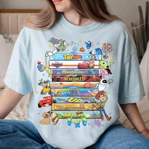 May include: Light blue t-shirt featuring a stack of colorful movie titles and characters from Disney Pixar films, including "Toy Story," "Cars," and "The Incredibles." The design includes characters like Buzz Lightyear, Woody, and Mike Wazowski.