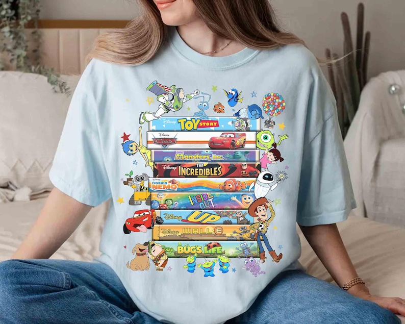 May include: Light blue t-shirt featuring a stack of colorful movie titles and characters from Disney Pixar films, including "Toy Story," "Cars," and "The Incredibles." The design includes characters like Buzz Lightyear, Woody, and Mike Wazowski.