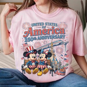 Może przedstawiać: Jasnor&oacute;żowa koszulka z Myszką Miki i Minnie w patriotycznych strojach, siedzących na ławce. Koszulka zawiera napis "United States of America 250th Anniversary" z fajerwerkami i pociągiem.