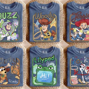 May include: Six blue t-shirts featuring Toy Story character graphics. The shirts display Buzz Lightyear, Woody, Jessie, Slinky Dog, a green frog named Lilypad, and Forky. Each design includes the character name in stylized text within a rectangular frame.