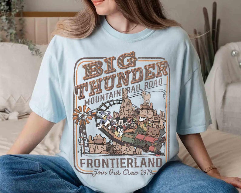 May include: Light blue t-shirt featuring a vintage-style graphic for Big Thunder Mountain Railroad. The design includes a train with Mickey Mouse and friends, a windmill, and the text "Frontierland Join Our Crew 1979."