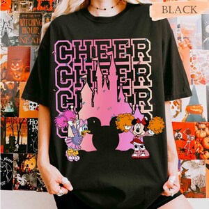 May include: Black t-shirt with the word "CHEER" stacked three times in a pink and white font. A pink castle silhouette is in the center. Cartoon characters Daisy Duck and Minnie Mouse are holding pom-poms.