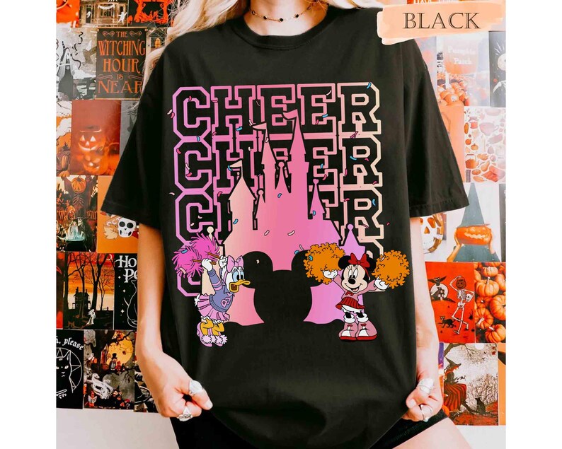May include: Black t-shirt with the word "CHEER" stacked three times in a pink and white font. A pink castle silhouette is in the center. Cartoon characters Daisy Duck and Minnie Mouse are holding pom-poms.