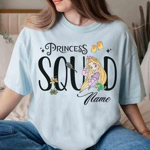 May include: Light blue t-shirt with the words "Princess Squad" and a cartoon illustration of a princess with long blonde hair. The shirt also features a flower, a frog, and two floating lanterns.