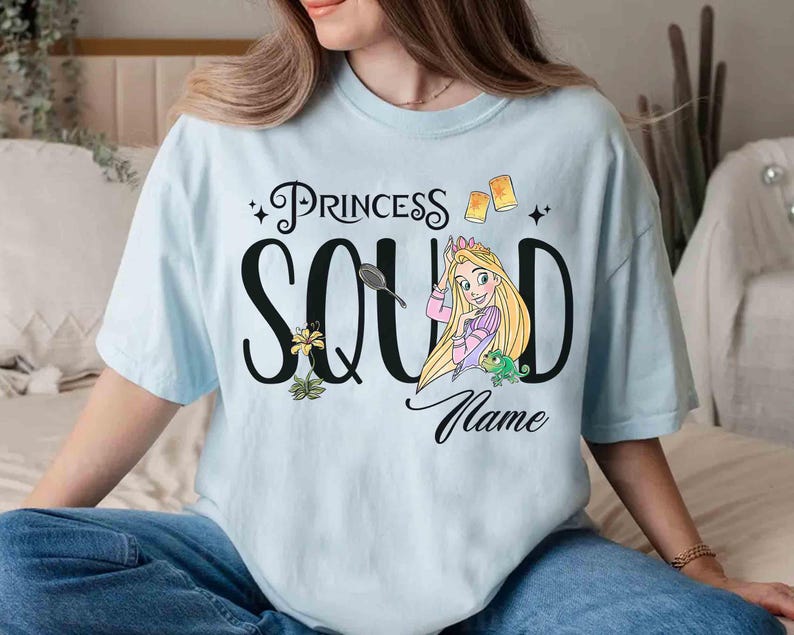 May include: Light blue t-shirt with the words "Princess Squad" and a cartoon illustration of a princess with long blonde hair. The shirt also features a flower, a frog, and two floating lanterns.