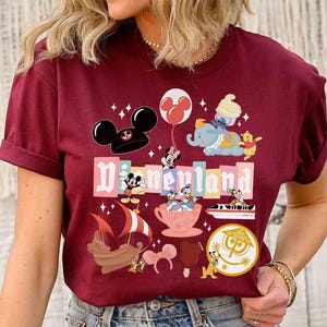 May include: Burgundy t-shirt featuring a Disneyland graphic with various Disney characters and icons. The design includes Mickey Mouse, Minnie Mouse, Dumbo, Winnie the Pooh, and other classic characters. The word "Disneyland" is prominently displayed in a colorful font.