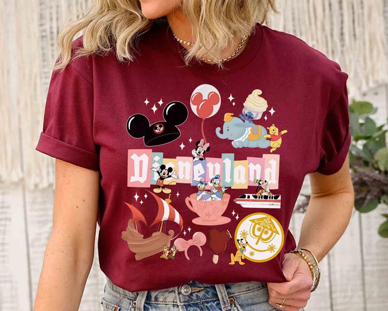 May include: Burgundy t-shirt featuring a Disneyland graphic with various Disney characters and icons. The design includes Mickey Mouse, Minnie Mouse, Dumbo, Winnie the Pooh, and other classic characters. The word "Disneyland" is prominently displayed in a colorful font.