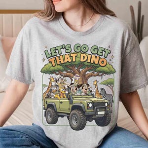 May include: A light gray t-shirt with the text "LET'S GO GET THAT DINO" above a cartoon safari scene. The scene includes Mickey and Minnie Mouse, a green jeep, giraffes, and a zebra.