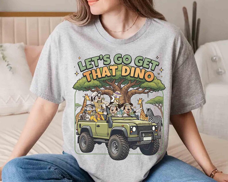 May include: A light gray t-shirt with the text "LET'S GO GET THAT DINO" above a cartoon safari scene. The scene includes Mickey and Minnie Mouse, a green jeep, giraffes, and a zebra.