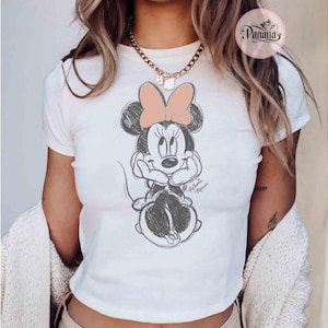 May include: White t-shirt with a black and white sketch of Minnie Mouse wearing a pink bow. The shirt has short sleeves and a crew neck.