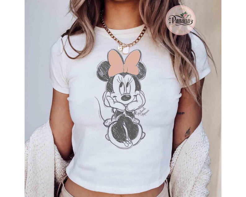 May include: White t-shirt with a black and white sketch of Minnie Mouse wearing a pink bow. The shirt has short sleeves and a crew neck.