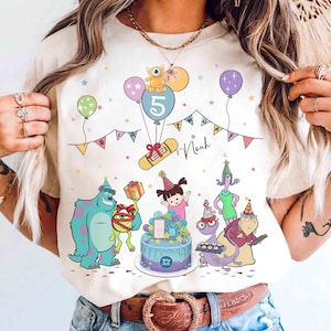 Puede incluir: Camiseta color crema con un diseño de cumpleaños colorido con personajes de la película Monstruos S.A. El diseño incluye globos, una tarta y el número 5. También se muestra el nombre "Noah".