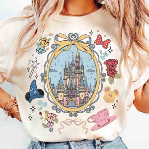May include: A white t-shirt featuring a colourful illustration of Cinderella's castle within a gold frame. The design incorporates flowers, stars, a magic wand, Mickey Mouse ears and a teacup.