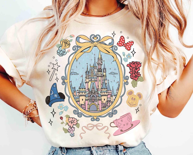May include: A white t-shirt featuring a colourful illustration of Cinderella's castle within a gold frame. The design incorporates flowers, stars, a magic wand, Mickey Mouse ears and a teacup.