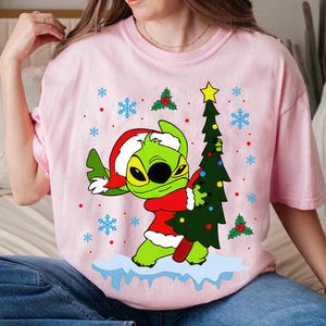 May include: Pink t-shirt featuring a green cartoon character in a Santa hat and red coat, holding a Christmas tree. The design includes snowflakes and holly berries. The shirt is a festive holiday item.