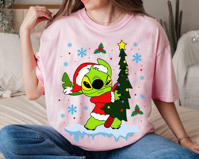 May include: Pink t-shirt featuring a green cartoon character in a Santa hat and red coat, holding a Christmas tree. The design includes snowflakes and holly berries. The shirt is a festive holiday item.