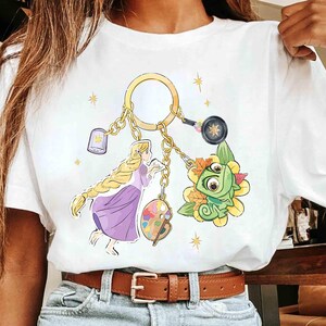 May include: White t-shirt featuring a gold keychain with Rapunzel, Pascal, and other themed charms. The design includes a lantern, a frying pan, and a palette. The shirt has a brown belt and blue jeans.