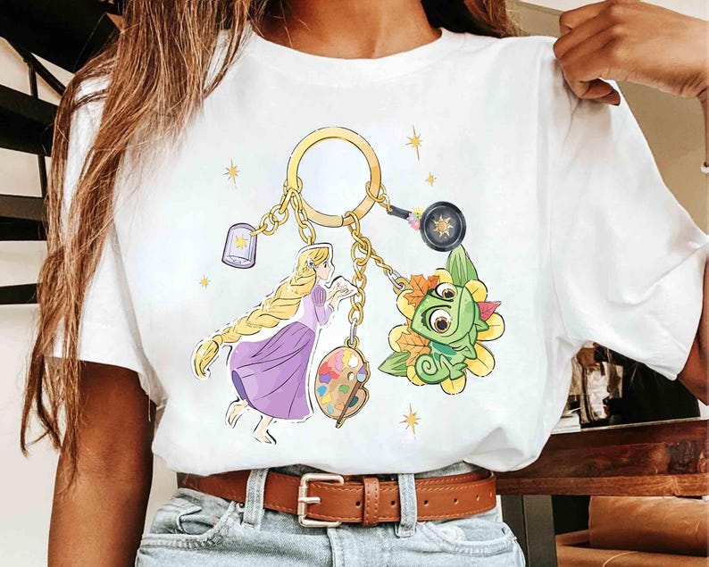 May include: White t-shirt featuring a gold keychain with Rapunzel, Pascal, and other themed charms. The design includes a lantern, a frying pan, and a palette. The shirt has a brown belt and blue jeans.