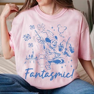 Puede incluir: Camiseta rosa claro con un dise&ntilde;o azul de Mickey Mouse como hechicero, sosteniendo una varita y cubos, con un castillo y fuegos artificiales. La palabra "Fantasmic!" est&aacute; escrita en azul debajo.