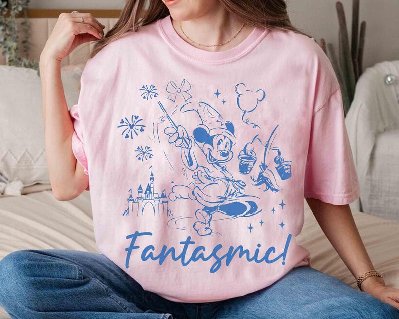 Puede incluir: Camiseta rosa claro con un dise&ntilde;o azul de Mickey Mouse como hechicero, sosteniendo una varita y cubos, con un castillo y fuegos artificiales. La palabra "Fantasmic!" est&aacute; escrita en azul debajo.