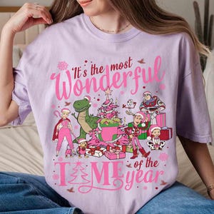 May include: A light purple t-shirt with a festive graphic. The design features the text "It's the most Wonderful Time of the Year" in pink, along with cartoon characters, a Christmas tree, and presents. The shirt is a relaxed fit.