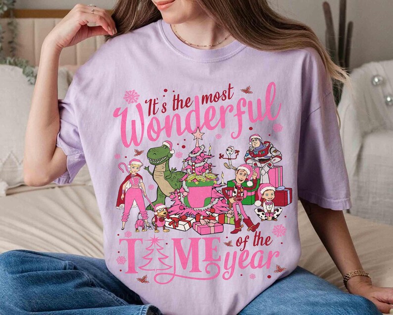 May include: A light purple t-shirt with a festive graphic. The design features the text "It's the most Wonderful Time of the Year" in pink, along with cartoon characters, a Christmas tree, and presents. The shirt is a relaxed fit.