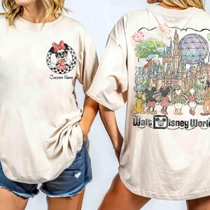 May include: Beige t-shirt featuring Minnie Mouse in a checkered circle with the text "Custom Name" on the front. The back of the shirt displays a colorful illustration of Walt Disney World with various characters and the words "Walt Disney World".