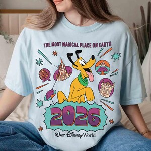 May include: Pale blue t-shirt with a Disney World design. It features Pluto, the text "THE MOST MAGICAL PLACE ON EARTH", and the year "2026". Various Disney park icons and the Walt Disney World logo are also present.