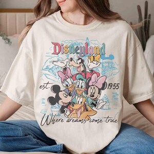 May include: Cream-colored t-shirt featuring a colorful Disneyland graphic with Mickey Mouse, Minnie Mouse, Donald Duck, Goofy, and Daisy Duck. The design includes the text "Disneyland est. 1955 Where dreams come true."