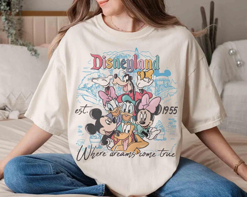 May include: Cream-colored t-shirt featuring a colorful Disneyland graphic with Mickey Mouse, Minnie Mouse, Donald Duck, Goofy, and Daisy Duck. The design includes the text "Disneyland est. 1955 Where dreams come true."